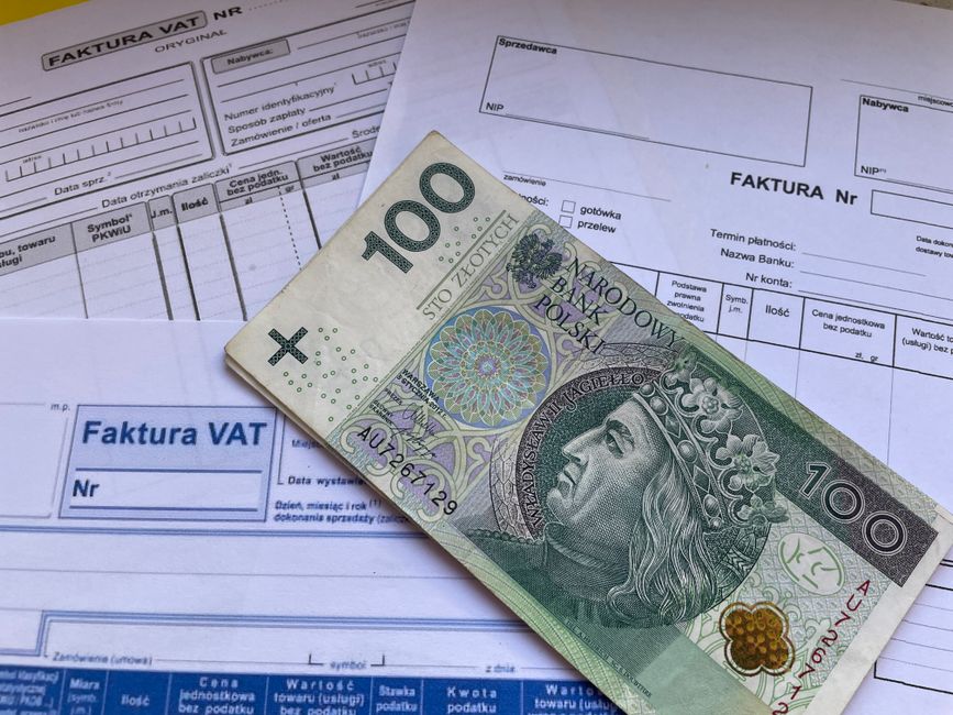 A close-up view of a 100 Polish złoty banknote placed diagonally over several invoices, including a VAT invoice, on a light-colored surface. The banknote features a portrait of a historical figure wi