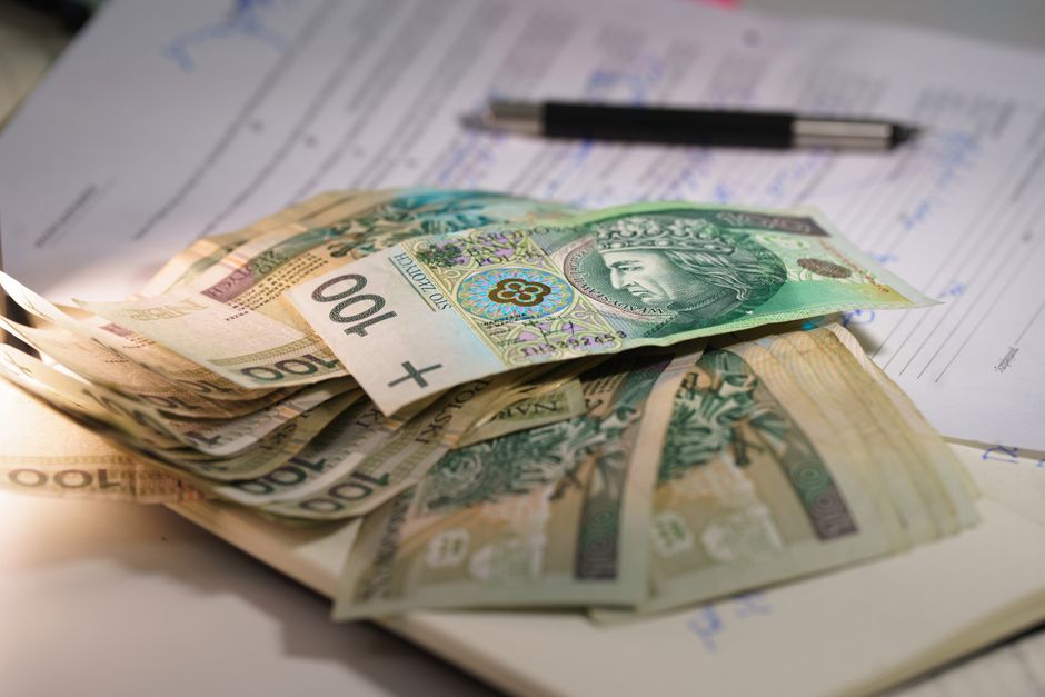 A close-up view of a 100 Polish złoty banknote placed diagonally over several invoices, including a VAT invoice, on a light-colored surface. The banknote features a portrait of a historical figure wi
