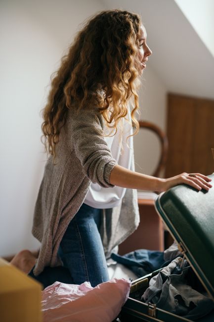 A man and woman inside a residential property during a home relocation, standing next to packing boxes and a partially open suitcase. The man, with long dark hair tied back, is smiling and gesturing t