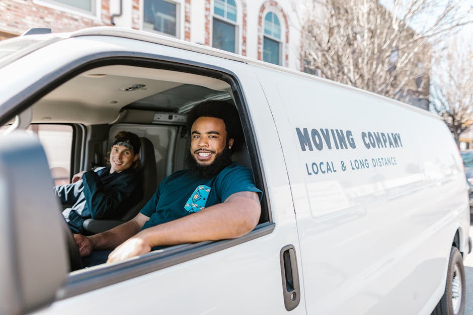 A white moving van parked outside a two-storey residential building with a brick and white facade. The van displays the words 'MOVING COMPANY' and 'LOCAL & LONG DISTANCE' on its side. Two men are pres