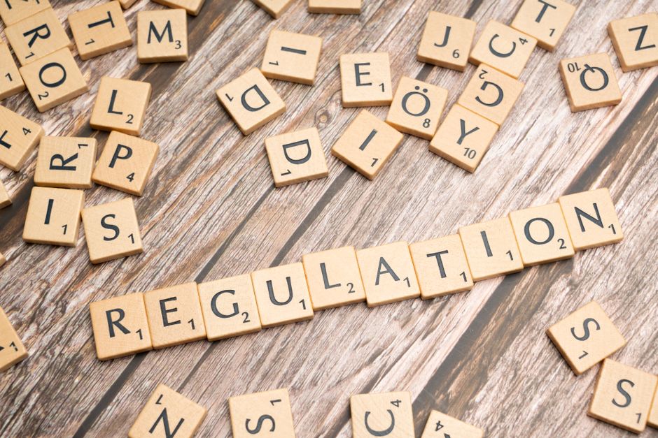 A collection of wooden Scrabble tiles scattered across a rustic wooden floor, with some tiles face-up and others face-down. Prominently, letters spell out the word 'REGULATION' in the center. The tile