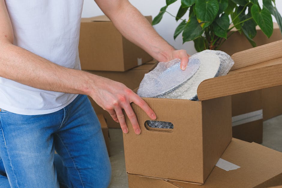 A person holding an open cardboard box with a red and white 'Fragile' sticker on the front, amidst a collection of packed moving boxes in a property setting. The individual’s hands are visible gripp