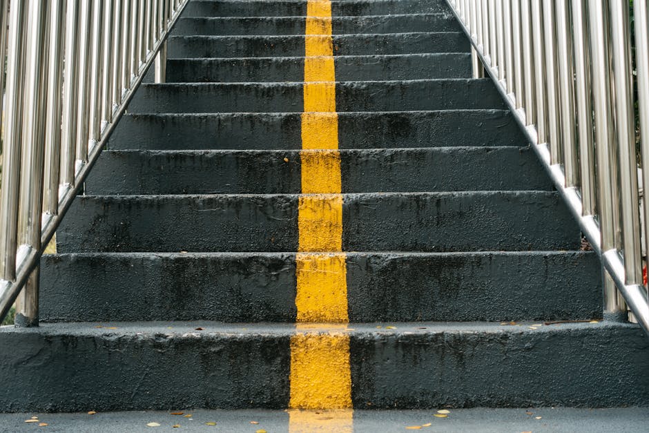 Indoor view of a staircase inside a building leading to an outdoor area, with brick walls on either side and metal handrails on both sides of the staircase. The stairs are made of dark-colored materia
