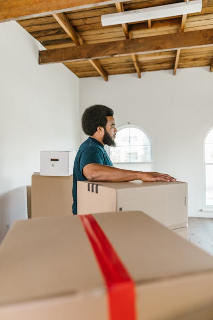 A man with dark skin, wearing a blue t-shirt, stands inside a bright, spacious room with a wooden beam ceiling and large arched windows, surrounded by stacked cardboard moving boxes of various sizes.